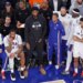 Philadelphia 76ers center Joel Embiid sits on the bench in plain clothes with teammates during a game.