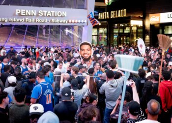 New York Knicks fans celebrating at Penn Station with a broom and a large cutout of Jalen Brunson's head.
