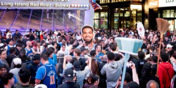 New York Knicks fans celebrating at Penn Station with a broom and a large cutout of Jalen Brunson's head.