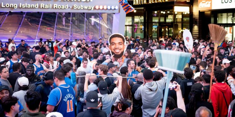 New York Knicks fans celebrating at Penn Station with a broom and a large cutout of Jalen Brunson's head.