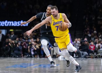 Los Angeles Lakers player in yellow jersey dribbling past a Brooklyn Nets player in black jersey.