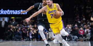 Los Angeles Lakers player in yellow jersey dribbling past a Brooklyn Nets player in black jersey.