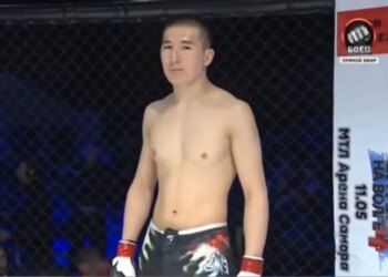 Medet Zheenaliev, shirtless and wearing fight shorts and gloves, standing in a fighting cage.