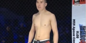 Medet Zheenaliev, shirtless and wearing fight shorts and gloves, standing in a fighting cage.