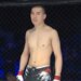 Medet Zheenaliev, shirtless and wearing fight shorts and gloves, standing in a fighting cage.
