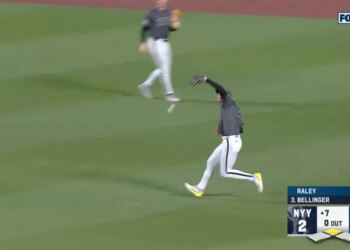 A baseball player in a gray and white uniform catches a ball on a green field as another player runs behind him.