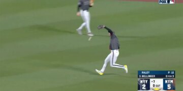 A baseball player in a gray and white uniform catches a ball on a green field as another player runs behind him.