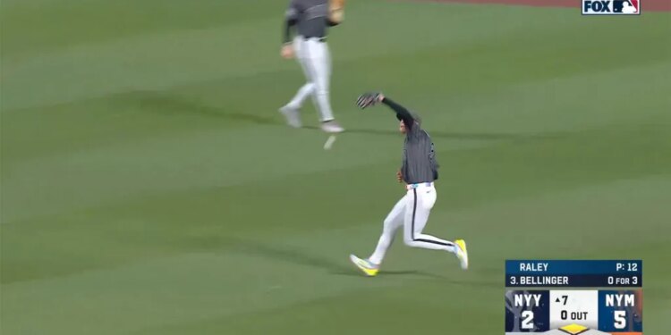 A baseball player in a gray and white uniform catches a ball on a green field as another player runs behind him.