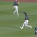 A baseball player in a gray and white uniform catches a ball on a green field as another player runs behind him.
