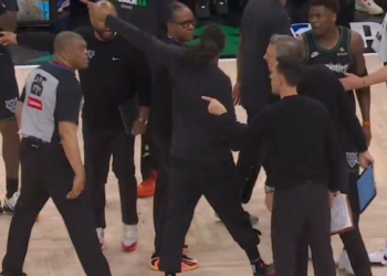 A woman in black clothing points to a referee, while several other people, including basketball players and staff, stand around them on a basketball court.