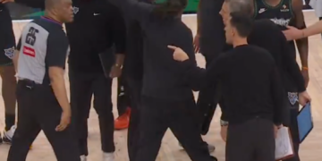 A woman in black clothing points to a referee, while several other people, including basketball players and staff, stand around them on a basketball court.