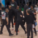 A woman in black clothing points to a referee, while several other people, including basketball players and staff, stand around them on a basketball court.