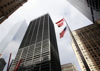 A low-angle photo looking up at tall buildings surrounding One Liberty Plaza in New York City, with an American flag and BBH flag flying between them.