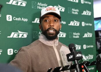 A man with a beard and a hat standing in front of a green banner with "JETS" and sponsor logos, speaking into a microphone.