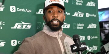A man with a beard and a hat standing in front of a green banner with "JETS" and sponsor logos, speaking into a microphone.
