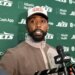 A man with a beard and a hat standing in front of a green banner with "JETS" and sponsor logos, speaking into a microphone.