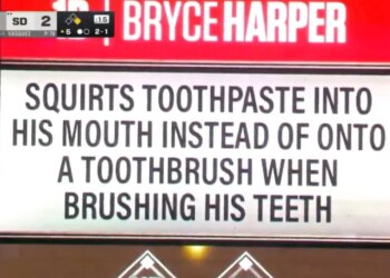 A scoreboard displays "BRYCE HARPER" in red and a white sign below it with black text that reads "SQUIRTS TOOTHPASTE INTO HIS MOUTH INSTEAD OF ONTO A TOOTHBRUSH WHEN BRUSHING HIS TEETH."