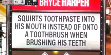 A scoreboard displays "BRYCE HARPER" in red and a white sign below it with black text that reads "SQUIRTS TOOTHPASTE INTO HIS MOUTH INSTEAD OF ONTO A TOOTHBRUSH WHEN BRUSHING HIS TEETH."
