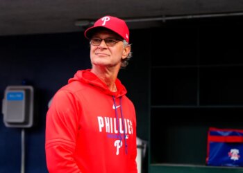 Don Mattingly in a Phillies baseball cap and red hoodie, standing in a dugout.