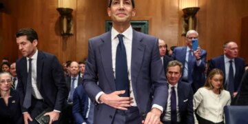 A man in a suit stands in a room with wood-paneled walls.