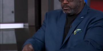 Shaquille O'Neal holding up tiny red and yellow shoes.