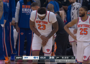 Mitchell Robinson, in a white New York Knicks jersey, clutches his right hand in pain.
