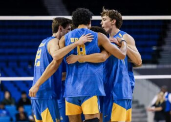 Four male volleyball players in blue and gold jerseys huddle together, arms around each other.