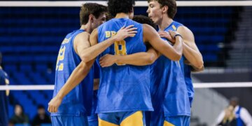 Four male volleyball players in blue and gold jerseys huddle together, arms around each other.