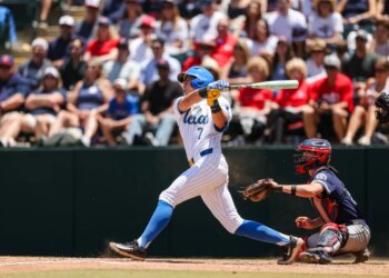 Roman Martin of UCLA batting a baseball with a catcher behind him.