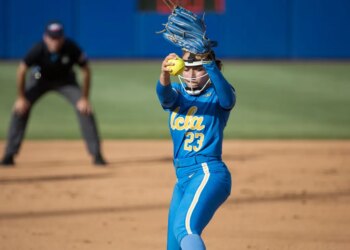 A UCLA softball pitcher in a blue uniform with the number 23 and the word "UCLA" on her chest, wearing a helmet and a blue glove, prepares to pitch a yellow softball with a blurry umpire in the background.