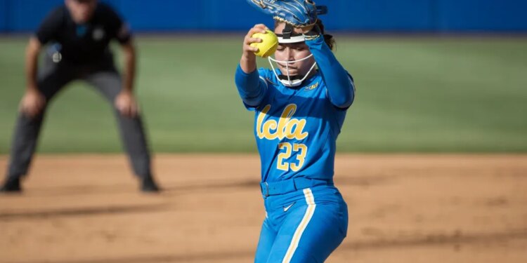 A UCLA softball pitcher in a blue uniform with the number 23 and the word "UCLA" on her chest, wearing a helmet and a blue glove, prepares to pitch a yellow softball with a blurry umpire in the background.