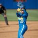 A UCLA softball pitcher in a blue uniform with the number 23 and the word "UCLA" on her chest, wearing a helmet and a blue glove, prepares to pitch a yellow softball with a blurry umpire in the background.