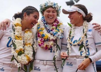 Three UCLA softball players in white uniforms, adorned with leis and flower crowns, laugh together on the field.