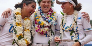 Three UCLA softball players in white uniforms, adorned with leis and flower crowns, laugh together on the field.