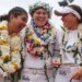 Three UCLA softball players in white uniforms, adorned with leis and flower crowns, laugh together on the field.