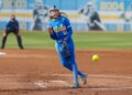 A UCLA softball pitcher in a blue uniform with yellow lettering pitches a neon yellow softball.