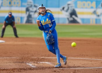 A UCLA softball pitcher in a blue uniform with yellow lettering pitches a neon yellow softball.