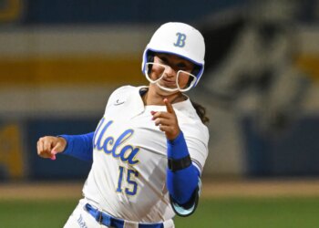 Jordan Woolery in a white and blue UCLA softball uniform, helmet and pink nails, pointing.