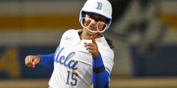 Jordan Woolery in a white and blue UCLA softball uniform, helmet and pink nails, pointing.