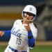 Jordan Woolery in a white and blue UCLA softball uniform, helmet and pink nails, pointing.