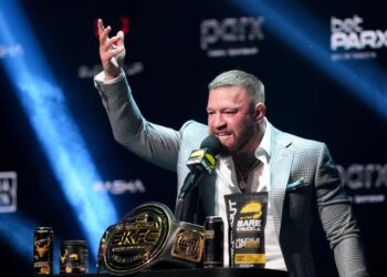 Conor McGregor speaking at a boxing news conference with a championship belt and products on a table in front of him.