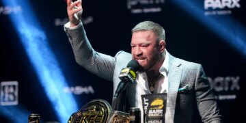 Conor McGregor speaking at a boxing news conference with a championship belt and products on a table in front of him.