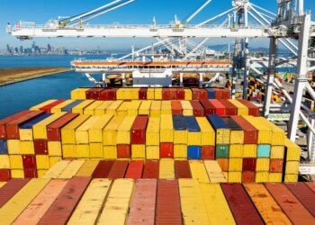 Cargo containers line a ship at the Port of Oakland with the city skyline in the background.