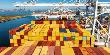 Cargo containers line a ship at the Port of Oakland with the city skyline in the background.