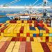 Cargo containers line a ship at the Port of Oakland with the city skyline in the background.