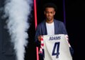Tyler Adams, a midfielder for the US men's national soccer team, holds up a jersey with "ADAMS" and the number 4 on the back, while being introduced during the 2026 FIFA World Cup roster announcement.