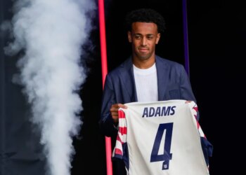 Tyler Adams, a midfielder for the US men's national soccer team, holds up a jersey with "ADAMS" and the number 4 on the back, while being introduced during the 2026 FIFA World Cup roster announcement.