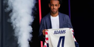 Tyler Adams, a midfielder for the US men's national soccer team, holds up a jersey with "ADAMS" and the number 4 on the back, while being introduced during the 2026 FIFA World Cup roster announcement.