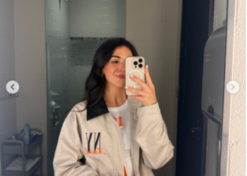 A woman takes a mirror selfie in a bathroom, wearing a beige jacket with black and orange details.
