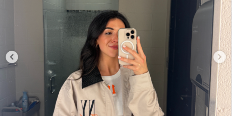 A woman takes a mirror selfie in a bathroom, wearing a beige jacket with black and orange details.